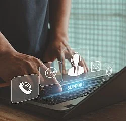 Eine Person nutzt einen Laptop, über dessen Tastatur holografische Icons für Kommunikation schweben. Zu sehen sind Symbole für Telefon, Chat, E-Mail und einen Support-Mitarbeiter mit Headset. Das Wort SUPPORT leuchtet in blauer Schrift, was Online-Kundenservice darstellt.