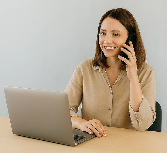 Eine lächelnde Frau sitzt an einem Schreibtisch vor einem Laptop und telefoniert mit einem Smartphone.